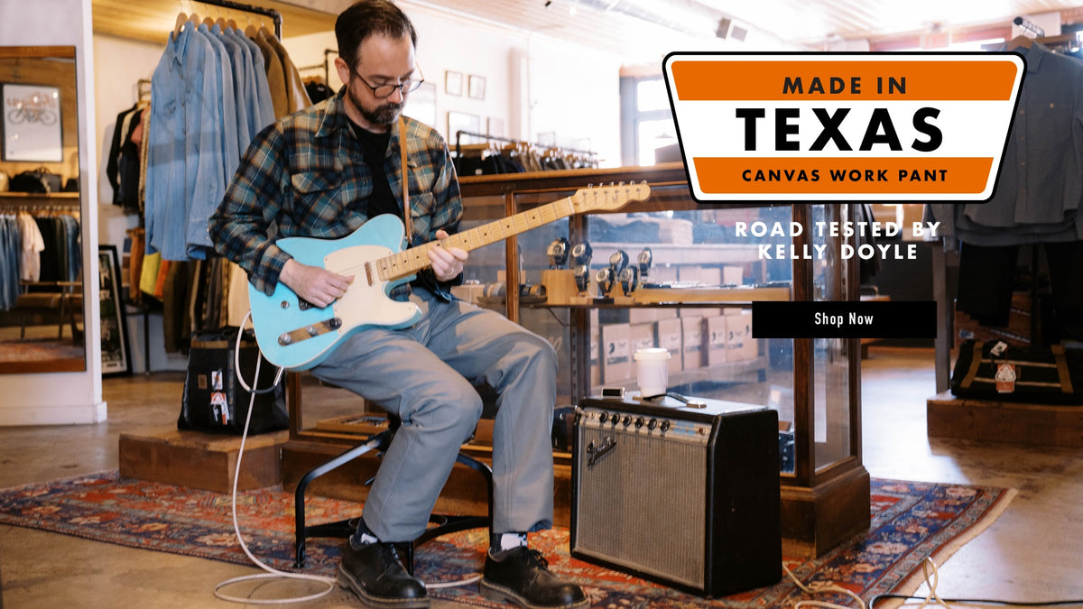 Man playing a blue electric guitar in a store with promotional text for 'Made in Texas' canvas work pants.